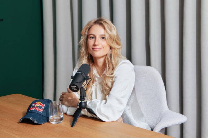 A podcast recording setup featuring a microphone on a wooden table. A navy blue cap with visible 'Red Bull' text is placed next to a clear glass. The setting includes a white chair and gray curtains in the background, creating a professional yet casual atmosphere.