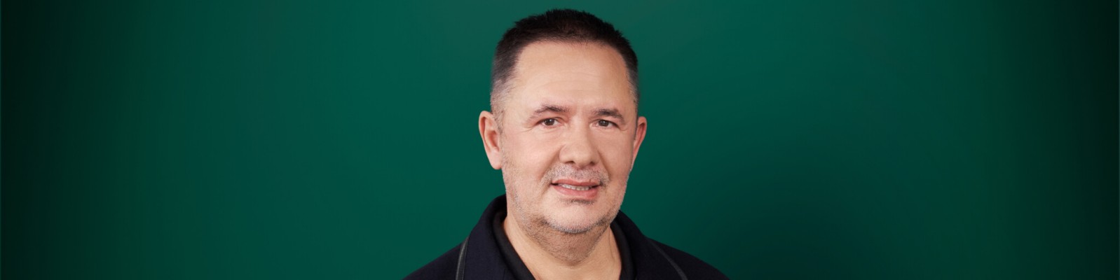 Samir Mane trägt ein schwarzes Hemd mit Kragen vor einem einfarbig grünen Hintergrund. Das Bild besticht durch eine schlichte und minimalistische Komposition ohne sichtbaren Text oder Logos. Die Atmosphäre ist neutral und professionell.