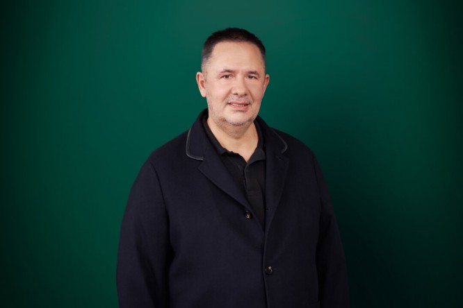 Samir Mane wearing a dark navy jacket and black shirt is standing against a solid green background. The setting is minimalistic, with no visible text or logos. The overall mood is formal and professional.