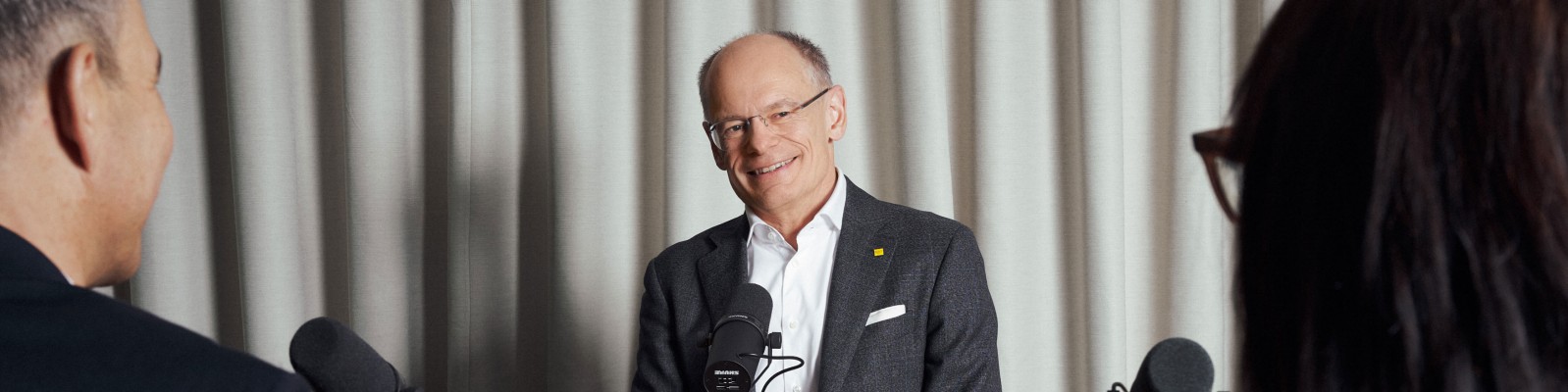 Personen sind in einem Interview-Setup mit Mikrofonen vor einem Vorhang-Hintergrund eingebunden. Die Szene vermittelt eine formelle und professionelle Umgebung.