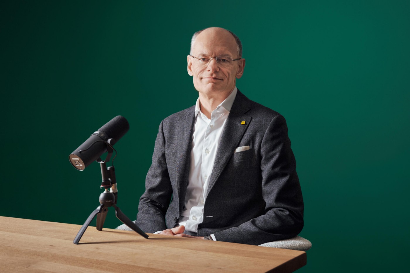 Eine Person in einem Anzug sitzt an einem Holztisch mit einem Mikrofon auf einem Ständer vor einem grünen Hintergrund. Repräsentiert eine Podcast- oder Interviewumgebung.