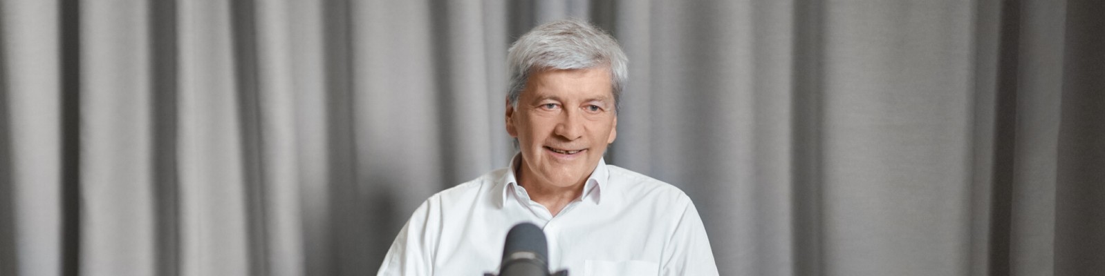 Dr. Johann Strobl wearing a white shirt is seated in front of a microphone. The setting features gray curtains as the backdrop, suggesting an indoor environment such as a studio or interview space. The microphone is prominently visible, indicating a recording or speaking activity.