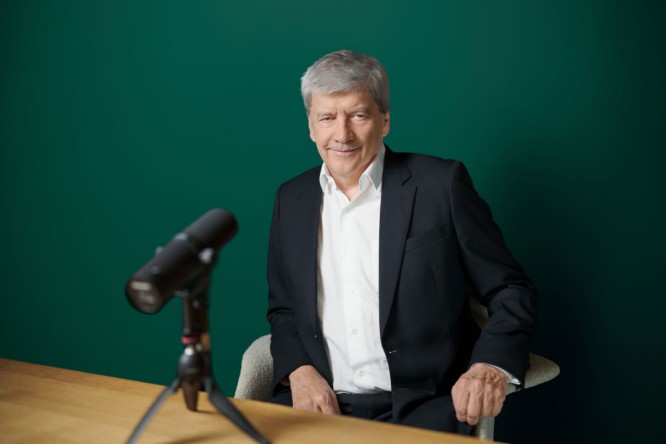 Dr. Strobl, gekleidet in einem schwarzen Anzug und weißen Hemd, sitzt an einem Holztisch mit einem professionellen Mikrofon auf einem Stativ vor sich. Der Hintergrund ist eine einfarbig grüne Wand, was eine klare und fokussierte Umgebung schafft. Das Bild deutet auf einen Podcast, ein Interview oder eine Aufnahmesession in einem professionellen Umfeld hin.