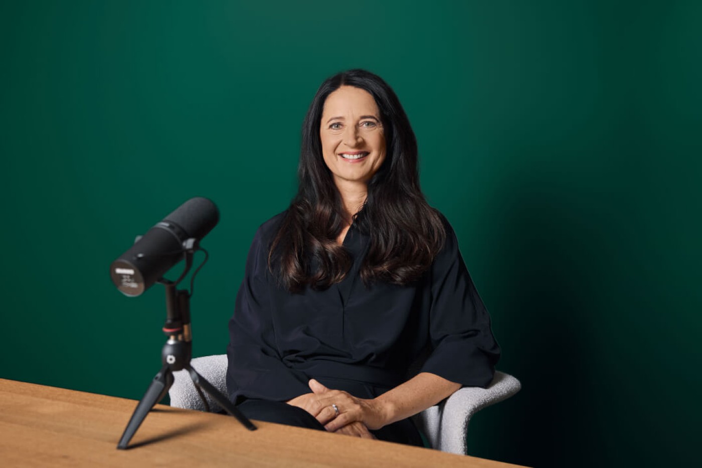 Marianne Heiß sitzt an einem Holztisch mit einem professionellen Mikrofon auf einem Stativ vor sich. Der Hintergrund ist eine einfarbig grüne Wand, die eine aufgeräumte und fokussierte Atmosphäre schafft. Das Mikrofon ist offenbar für Podcast- oder Aufnahmezwecke gedacht, und die Gesamtstimmung wirkt professionell und minimalistisch.