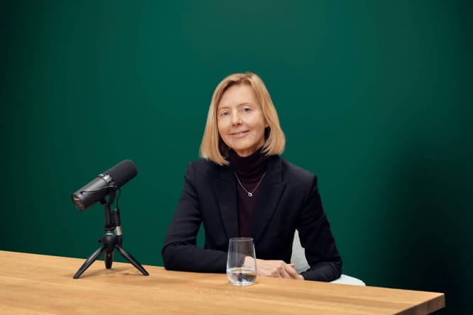 Marianne Janik sitzt an einem Holztisch in einem modernen Studio. Sie trägt einen dunklen Blazer und einen Rollkragenpullover. Links von ihr steht ein Mikrofon auf einem Stativ, vor ihr ein klares Glas Wasser. Der einfarbig grüne Hintergrund erzeugt eine professionelle und minimalistische Atmosphäre. Im Bild sind weder Text noch Zahlen zu sehen.