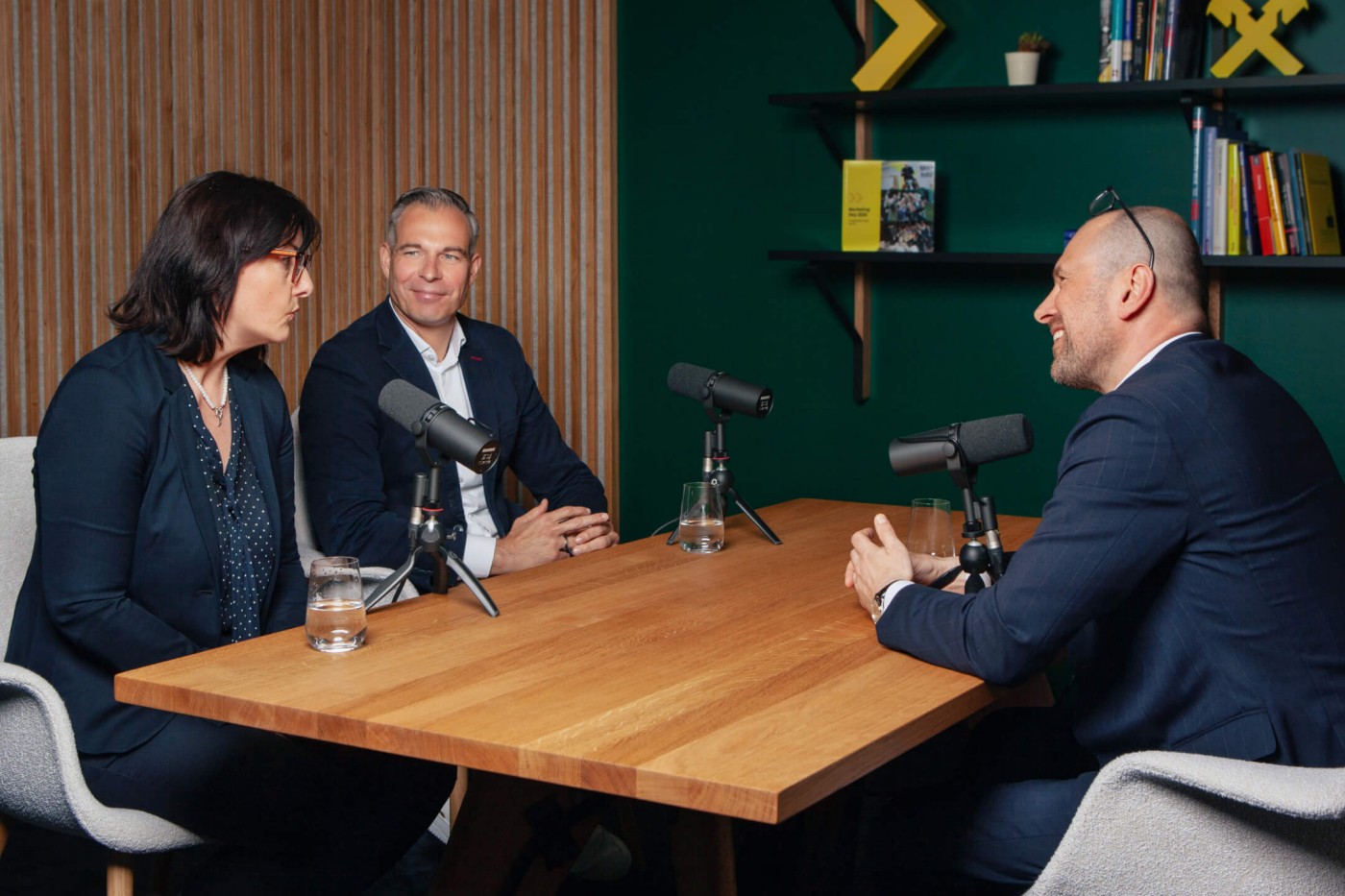 Drei Geschäftsleute in Businesskleidung sitzen an einem Tisch mit Mikrofonen und Wassergläsern und nehmen einen Podcast in einem modernen Büro auf.