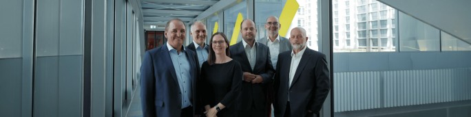 A group of six professionally dressed adults stands together in a modern indoor corridor. The people appear to be business colleagues posing for a group portrait, with glass walls and metal architectural elements surrounding them. Bright yellow diagonal shapes on the wall and a cityscape of tall buildings outside the windows add a contemporary urban feel. The overall mood is professional and confident, suggesting corporate leadership or executive teamwork.