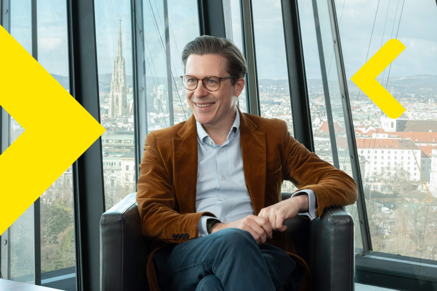 Björn Zaksek sitzt in einem braunen Blazer und einem hellblauen Hemd in einem modernen Sessel vor großen Glasfenstern. Im Hintergrund bietet sich ein Panoramablick auf die Stadt mit einem markanten Kirchturm. Die Szenerie wirkt wie ein Hochhaus oder eine Aussichtsplattform; geometrische gelbe Grafiken überlagern das Bild. Die Stimmung ist professionell und urban, die kühle Farbpalette wird durch die Kleidung des Mannes zusätzlich betont.