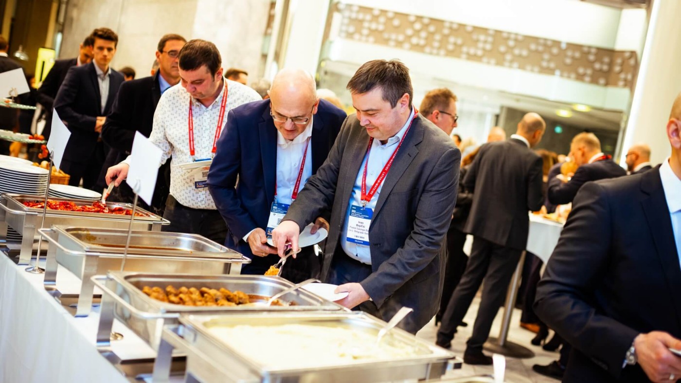 Eine Gruppe von Geschäftsleuten in Anzügen bedient sich an einem Buffet bei einer Konferenz, im Hintergrund viele weitere Personen.