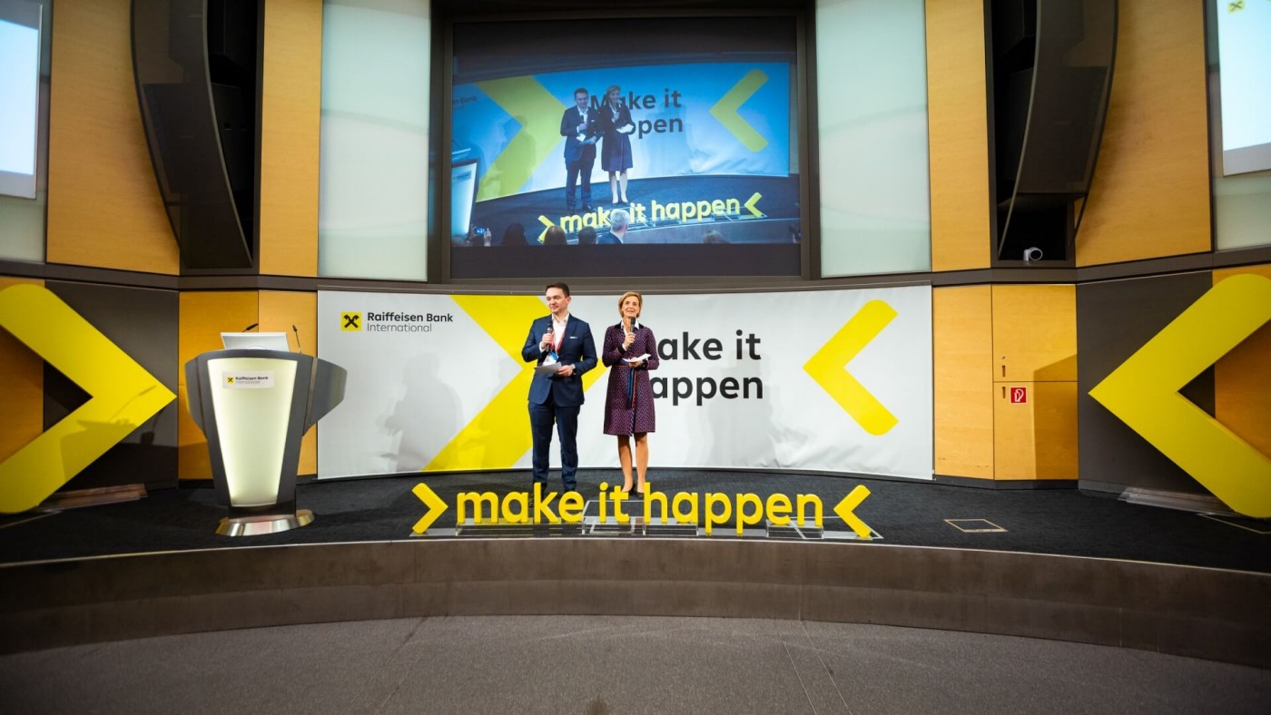 Zwei Referenten stehen auf der Bühne vor einem Hintergrund mit dem Slogan "make it happen" und Branding für Raiffeisen Bank International.