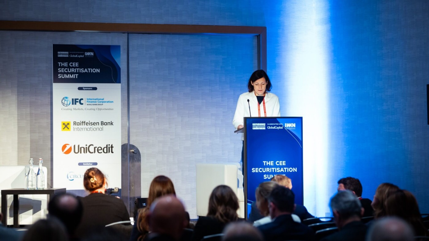 Eine Frau steht an einem Rednerpult und spricht vor einem Publikum beim CEE Securitisation Summit, mit großem Bildschirm und Sponsorenlogos im Hintergrund.