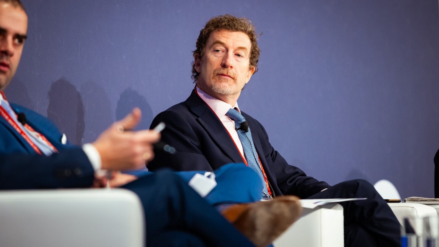 Zwei Männer in Anzügen sitzen auf der Bühne vor einem dunklen Hintergrund und führen während einer Konferenz eine Diskussion.