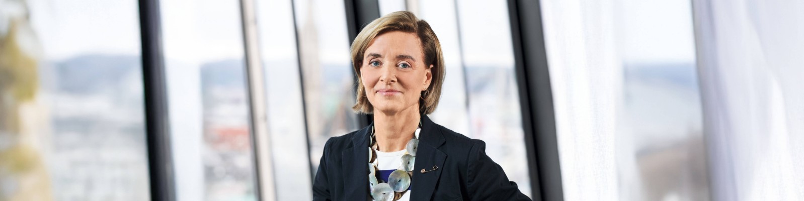 A person stands confidently in a modern office environment, wearing a dark blazer and a statement necklace. Large windows in the background offer a view of the cityscape.