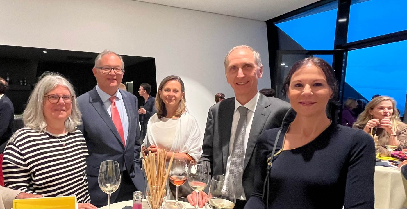 Eine Gruppe von Personen in formeller Kleidung ist bei einer gesellschaftlichen Veranstaltung versammelt. Die Szenerie spielt sich in Innenräumen ab, große Fenster bieten einen Blick in die Dämmerung. Auf dem Tisch stehen Weingläser und Häppchen, darunter Brotstangen. Die Atmosphäre wirkt stilvoll und entspannt.