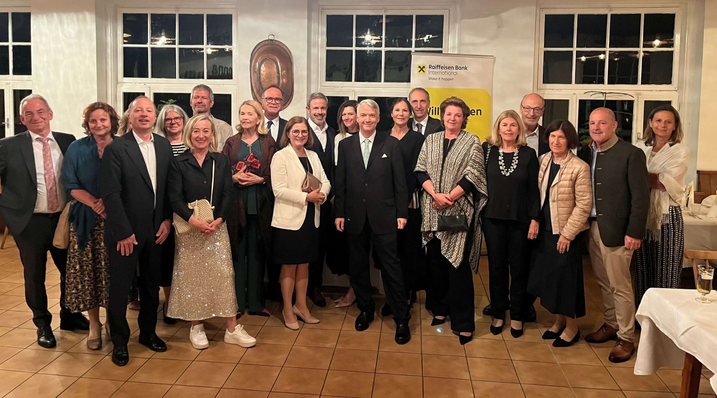 Eine Gruppe von Personen in formeller Kleidung posiert bei einer Veranstaltung in Innenräumen. Der Raum ist gut beleuchtet, mit großen Fenstern und Holzboden ausgestattet. Im Hintergrund ist ein Raiffeisen-Bank-Banner zu sehen, was auf eine Sponsoring-Partnerschaft oder Zugehörigkeit hinweist. Die Stimmung ist professionell und feierlich.