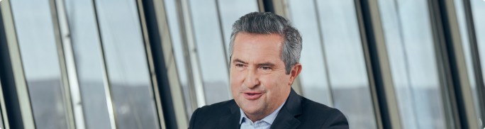 Rainer Schnabl is seated in a contemporary office environment. The person is dressed in a dark suit jacket and light shirt, suggesting a formal corporate setting. Large vertical glass windows in the background reveal an urban or industrial landscape. The overall scene conveys professionalism and a modern workplace atmosphere.
