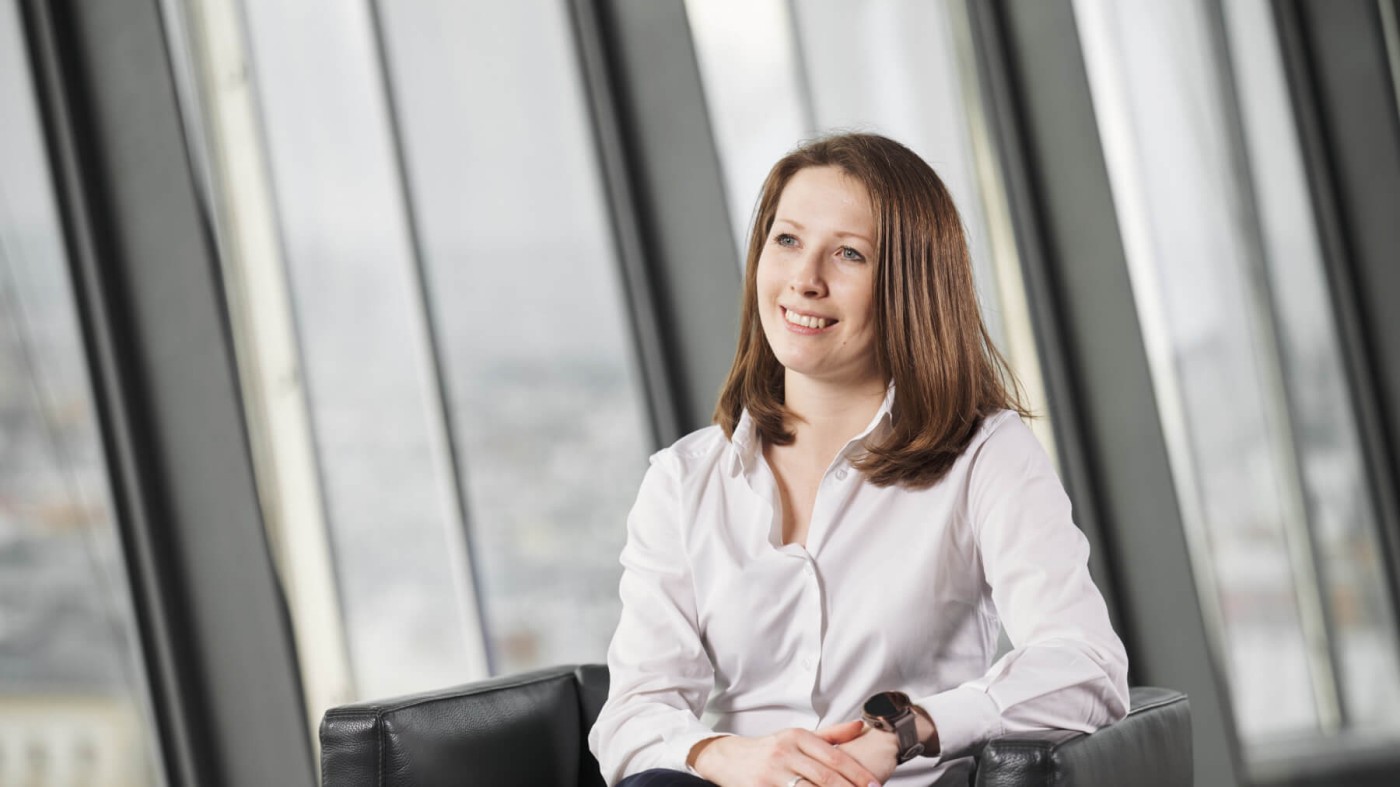 Evgeniya Sharkova wearing a white button-up shirt is seated on a black leather chair in a contemporary office setting. Large glass windows in the background reveal an urban cityscape. The atmosphere is professional and minimalistic, with neutral tones dominating the palette.