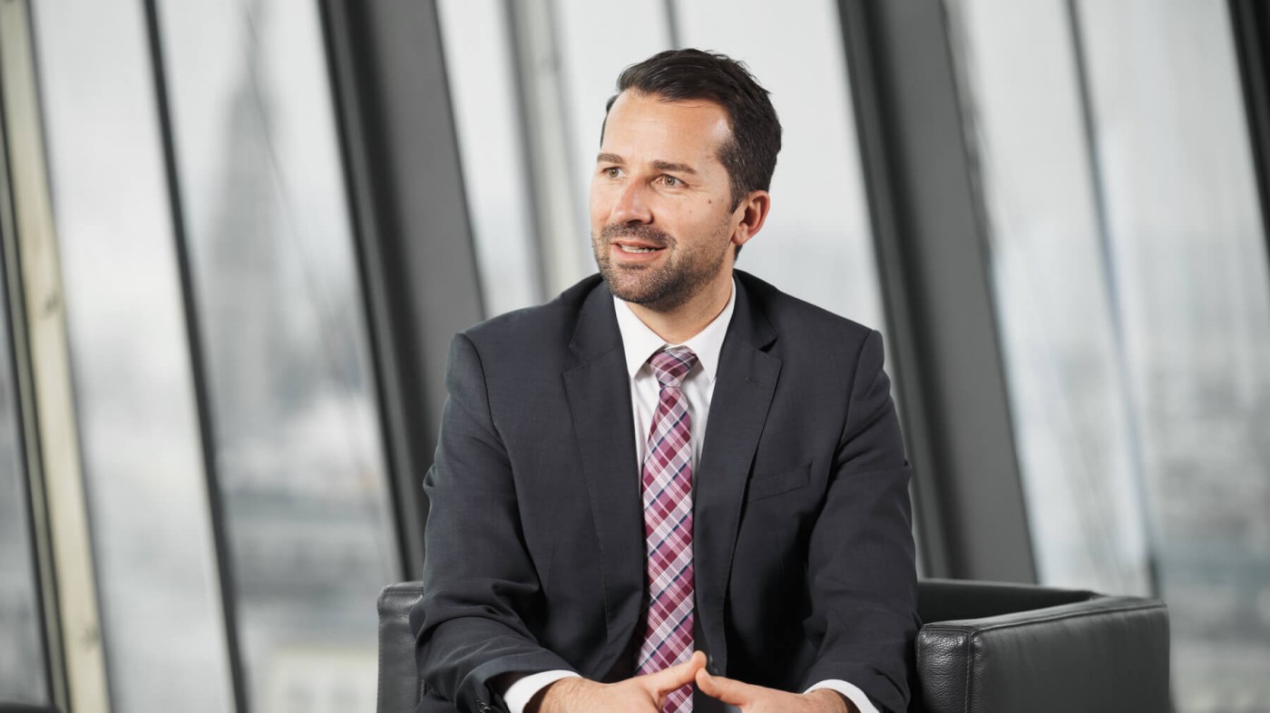 Sanin Merdžan wearing a dark suit and patterned tie is seated in a modern office setting. The background features large glass windows with an urban cityscape visible. The atmosphere conveys professionalism and corporate style.