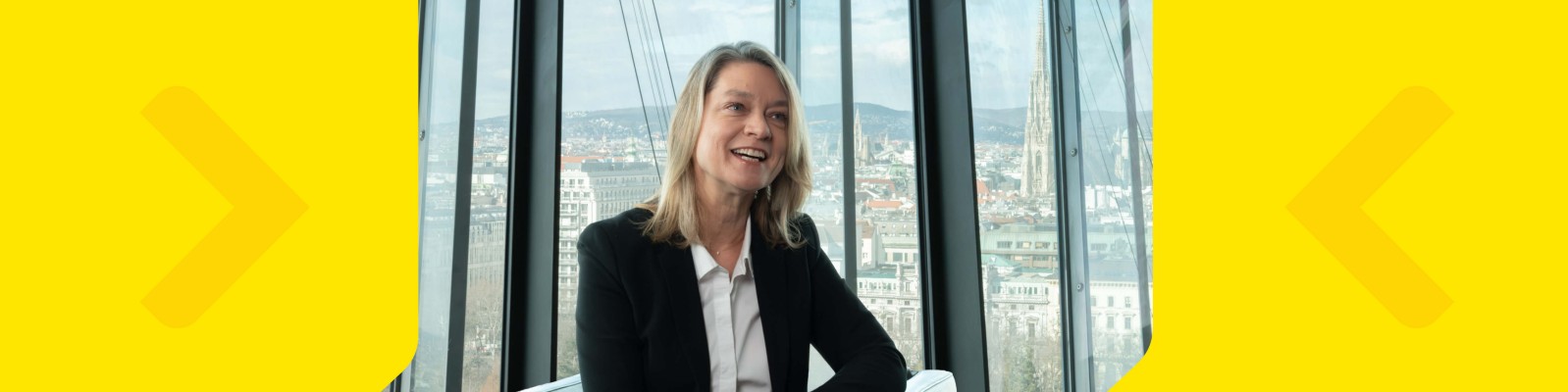 A woman in professional attire sits in a contemporary office space with large glass windows overlooking a cityscape. The setting features bright yellow graphic elements framing the image, adding a modern and energetic touch. The woman appears to be in her 30s or 40s, dressed in a black blazer and white shirt, suggesting a business or corporate environment. The overall mood is professional and dynamic, with natural daylight illuminating the scene.
