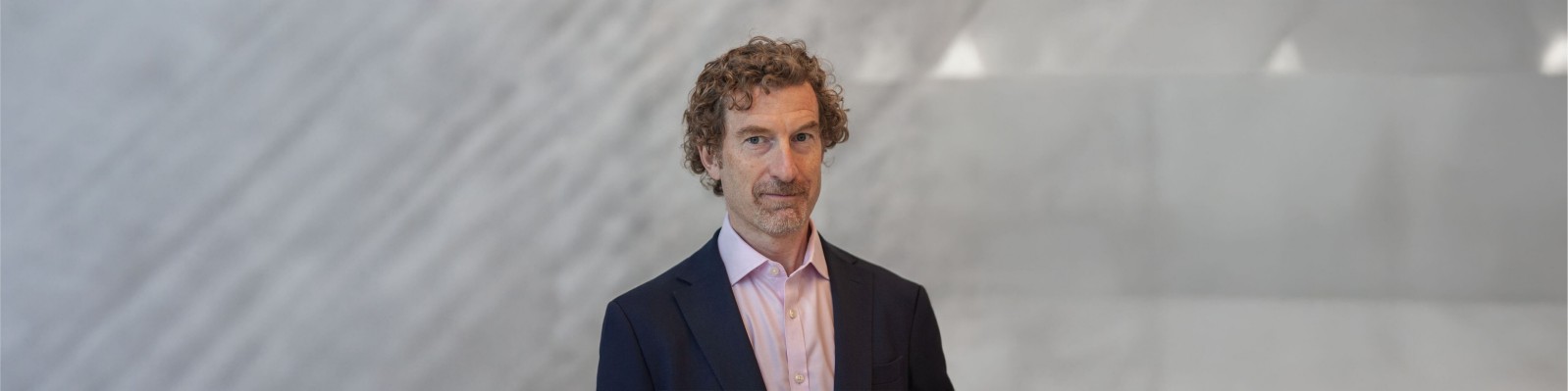 Martin Blum dressed in a dark suit jacket and light pink shirt, showcasing curly hair. The background is a soft, blurred gray pattern.
