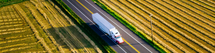 A white semi truck travels along a straight, two-lane highway bordered by golden agricultural fields. The scene is captured from above, highlighting the geometric patterns of the farmland and the long shadow cast by the vehicle. The image conveys a sense of movement, open space, and rural industry under clear daylight.