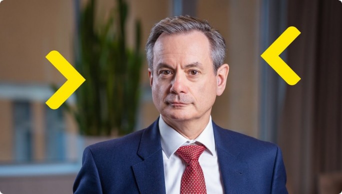 Laszlo Havas professionally dressed, wearing a navy blue suit, white shirt, and red tie is seated indoors. The background features a blurred office setting with a plant and large windows. Yellow arrow graphics are overlaid on the image, adding a modern design element.