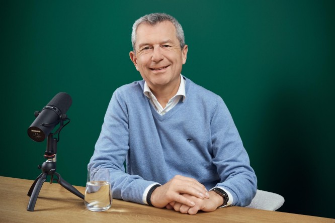 Kurt Svoboda is speaking into a studio microphone while sitting at a wooden desk. The person wears a light blue sweater over a collared shirt, with hands resting calmly on the table. A glass of water is placed beside the microphone, suggesting a relaxed yet focused recording environment. The background is a solid green color, emphasizing the subject and podcast setup.