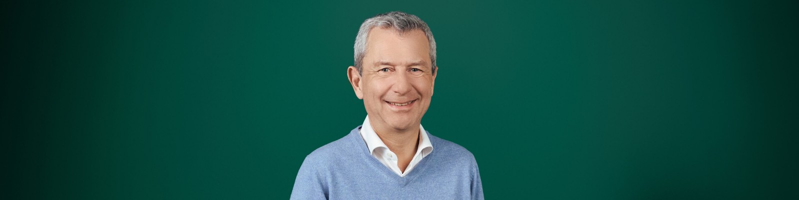 Kurt Svoboda with short gray hair is posed in front of a solid green studio backdrop. Their face is intentionally blurred for anonymity and privacy. They are wearing a light blue V-neck sweater layered over a white collared shirt. The image has a clean, minimal composition suitable for professional or corporate use.