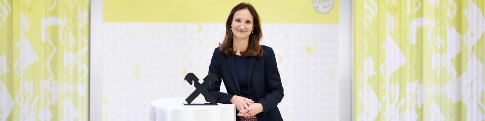 Eine professionell gekleidete Frau steht bei einer Preisverleihung hinter einem kleinen runden Rednerpult. Sie trägt einen dunklen Blazer und stützt die Hände neben einer stilisierten schwarzen Trophäe auf das Pult. Im Hintergrund ist eine leuchtend gelb-weiße Grafikwand mit dezenten Mustern und Textelementen zu sehen. Die Szene wirkt wie eine formelle Auszeichnungsveranstaltung eines Unternehmens oder einer Branche.