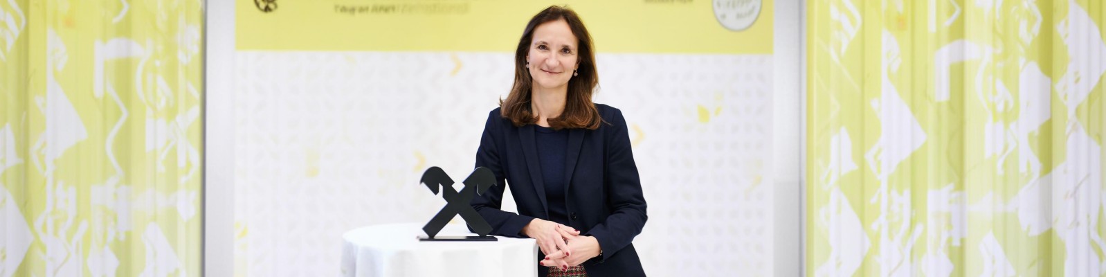 Eine professionell gekleidete Frau steht bei einer Preisverleihung hinter einem kleinen runden Rednerpult. Sie trägt einen dunklen Blazer und stützt die Hände neben einer stilisierten schwarzen Trophäe auf das Pult. Im Hintergrund ist eine leuchtend gelb-weiße Grafikwand mit dezenten Mustern und Textelementen zu sehen. Die Szene wirkt wie eine formelle Auszeichnungsveranstaltung eines Unternehmens oder einer Branche.
