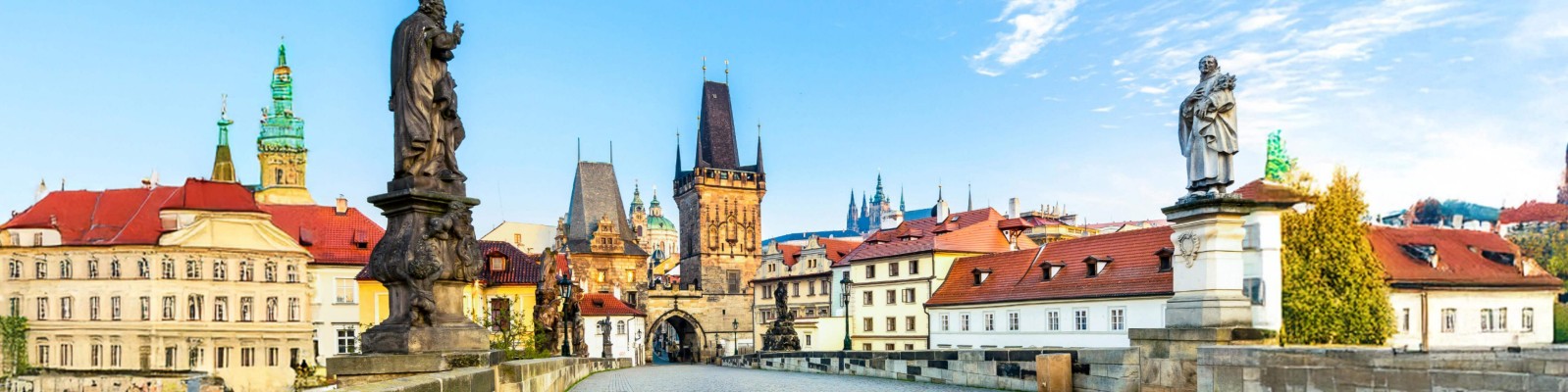A scenic view of Prague’s historic cityscape featuring statues on a bridge, red-roofed buildings, and iconic towers under a blue sky.