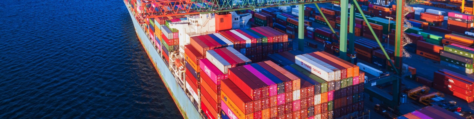 Eine Luftaufnahme eines großen Containerschiffs, das mit bunten Versandcontainern beladen und im Hafen liegt, umgeben von blauem Wasser. Das Bild fängt die lebendigen Farben und die geschäftige Atmosphäre der maritimen Logistik ein.