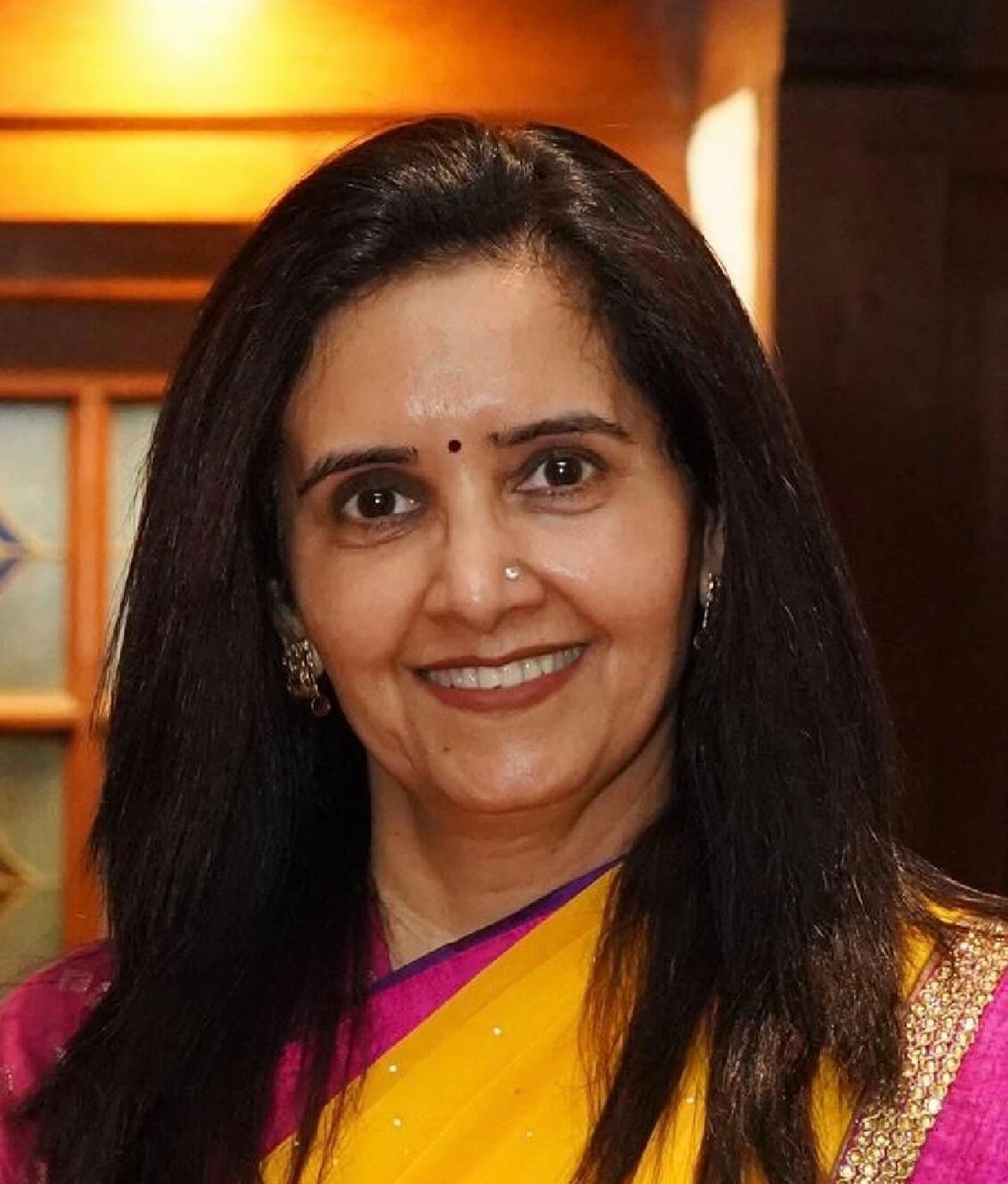 A woman wearing a bright yellow sari with a pink border stands indoors. The sari features subtle sparkling embellishments and a gold trim along the edge. Her long dark hair is worn straight over her shoulders. The background shows warm-toned wooden panels and a softly lit interior setting.