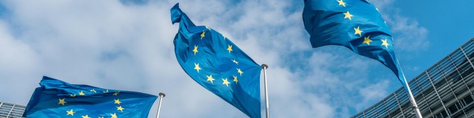 Three European Union flags are prominently displayed, waving against a backdrop of a partly cloudy blue sky. The flags feature a circle of yellow stars on a blue field, symbolizing unity. A modern building structure is partially visible in the background, adding an urban context.