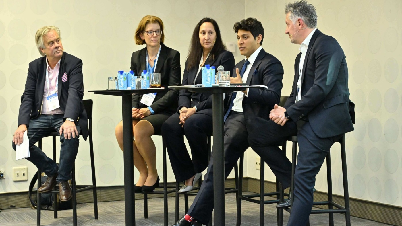 Eine Gruppe von Fachleuten sitzt an einem Hochtisch während einer Podiumsdiskussion im Geschäftsbereich. Die Umgebung erinnert an einen Konferenzraum mit neutralem Dekor. Auf dem Tisch sind Wasserflaschen deutlich sichtbar platziert, und die Teilnehmenden sind formell gekleidet. Die Atmosphäre wirkt professionell und von Zusammenarbeit geprägt.