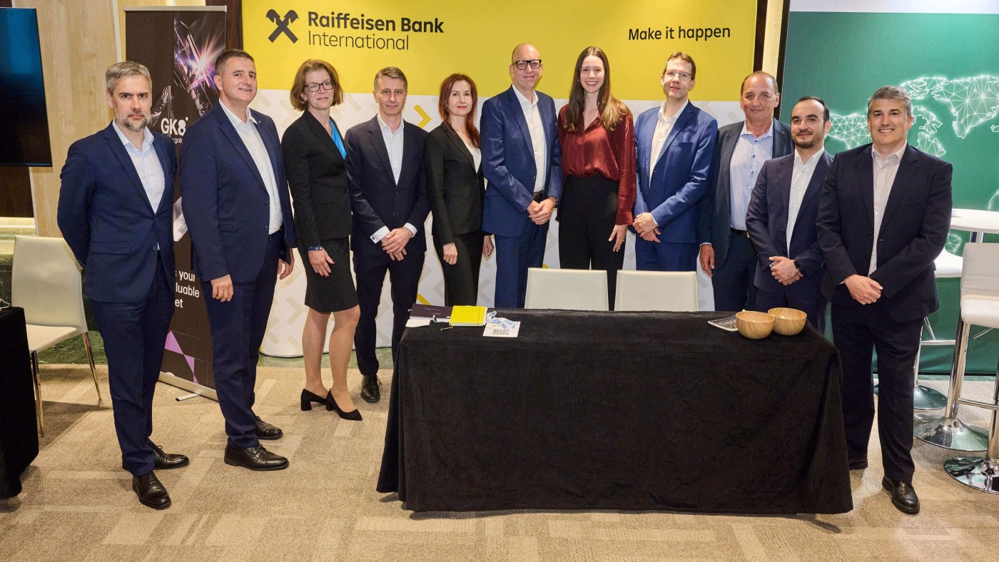 Eine Gruppe von Fachleuten in formeller Kleidung posiert vor einem Banner der Raiffeisen Bank International mit dem Slogan „Make it happen“. Die Szene spielt sich offenbar bei einer Firmenveranstaltung oder Konferenz ab; im Vordergrund steht ein Tisch, im Hintergrund sind weitere Banner zu sehen. Das Bild zeigt eine Mischung aus Männern und Frauen in Businesskleidung und vermittelt eine professionelle, kooperative Atmosphäre.