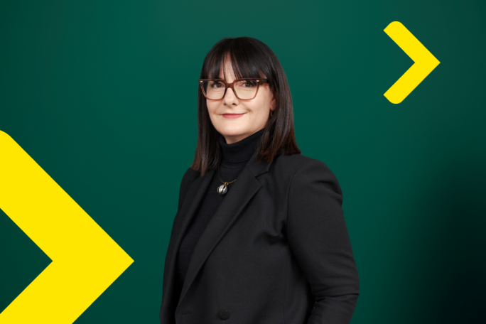 Elitza Kavrakova dressed in a black blazer and turtleneck stands against a dark green backdrop. The setting features bold yellow arrow graphics, adding a modern and dynamic visual element. The overall mood is professional and contemporary, with a focus on business attire and clean design. No visible text or numbers are present in the image.