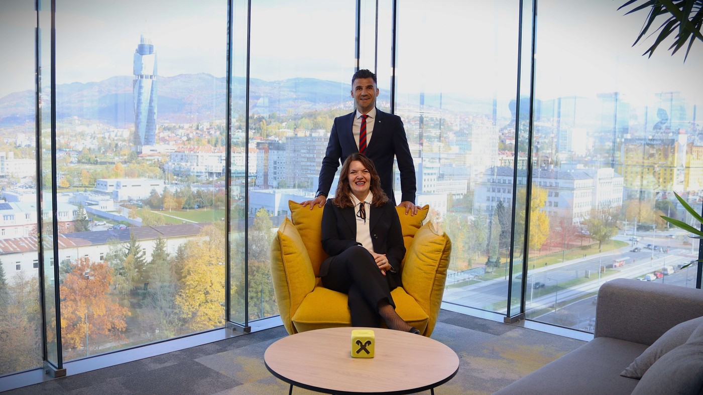 Zwei Geschäftsleute in formeller Kleidung befinden sich in einem modernen Büro mit großen Glasfenstern, die den Blick auf die Stadt und die umliegende Natur freigeben. Einer sitzt auf einem leuchtend gelben Stuhl, der andere steht hinter ihm. Im Vordergrund befindet sich ein kleiner Tisch mit einem Würfel, auf dem das Raiffeisen-Logo deutlich zu sehen ist. Im Hintergrund sind ein hohes Gebäude und herbstlich gefärbte Bäume zu erkennen.