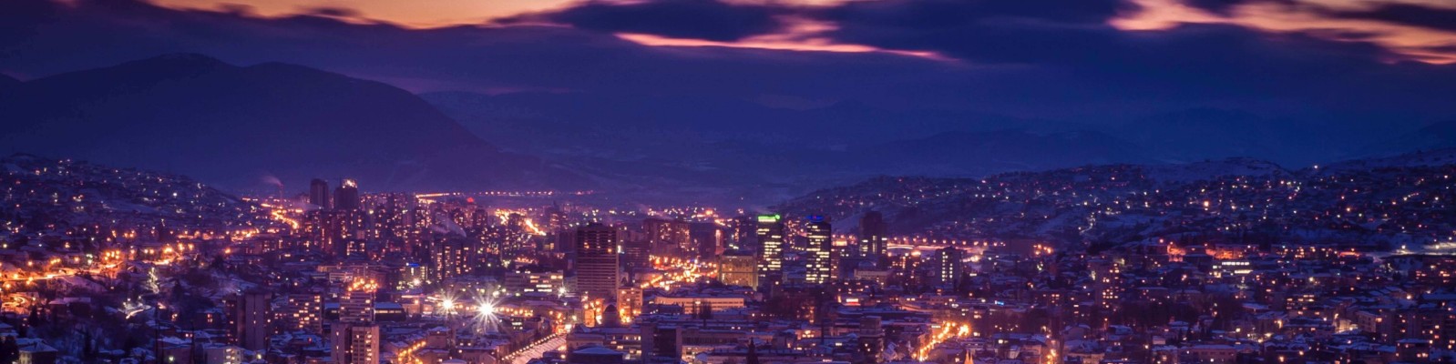 Ein Panoramablick auf eine Stadt, die in der Abenddämmerung von leuchtenden Lichtern erhellt wird. Im Hintergrund erhebt sich eine Bergkulisse unter einem dramatischen, violett-orangenen Himmel. Die Stadtlandschaft präsentiert Gebäude und Straßen, die im Abendlicht erstrahlen und eine zugleich ruhige und lebendige Atmosphäre schaffen.