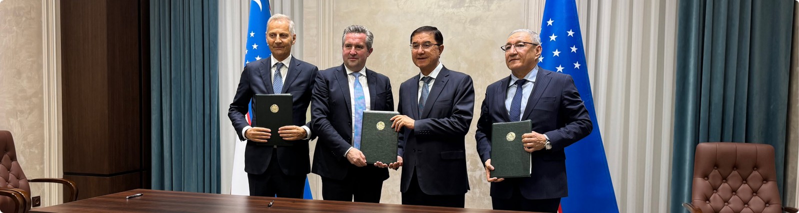 Vier formell gekleidete Männer stehen hinter einem großen Konferenztisch und halten dunkelgrüne Mappen in den Händen, was auf eine unterzeichnete Vereinbarung oder Partnerschaft hindeutet. Sie befinden sich zwischen zwei blauen Nationalflaggen mit weißen und grünen Streifen und weißen Sternen. Der Raum wirkt wie ein offizieller Regierungs- oder Firmenbesprechungsraum mit Holzvertäfelung und Ledersesseln. Die Atmosphäre ist formell und professionell und unterstreicht Diplomatie, Kooperation und erfolgreiche Verhandlungen.