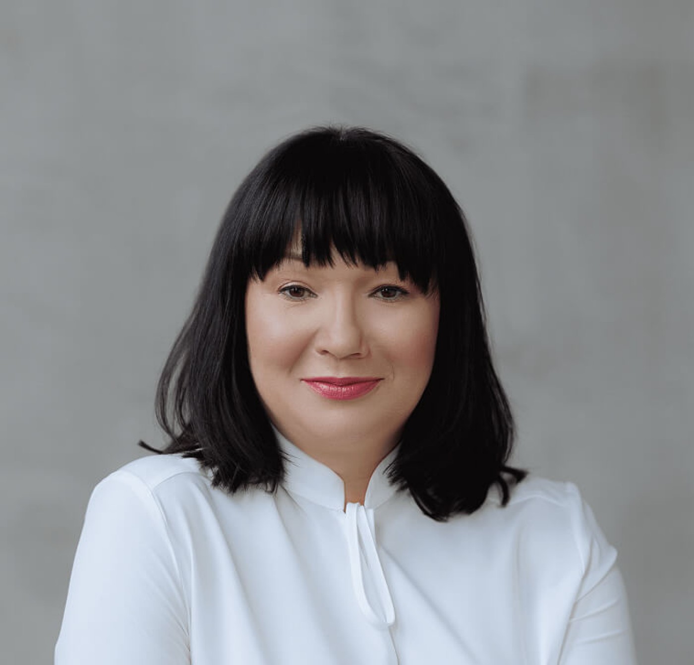A person with shoulder-length black hair, wearing a white blouse, set against a neutral gray background. The image conveys a formal and professional appearance.