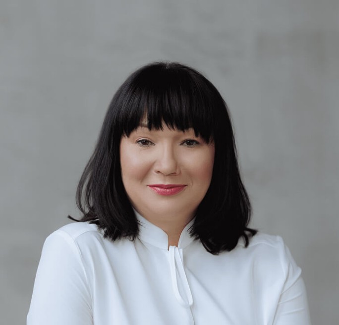 A person with shoulder-length black hair, wearing a white blouse, set against a neutral gray background. The image conveys a formal and professional appearance.