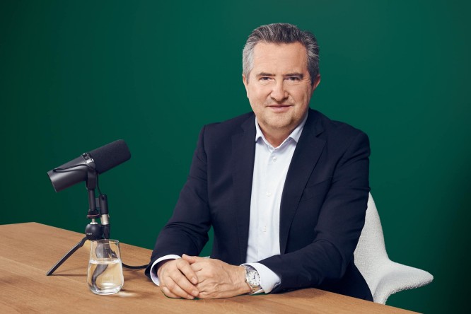 Rainer Schnabl in a dark suit sits at a wooden table in front of a studio microphone. He faces the camera with a confident, composed expression against a solid green background. A glass of water and the microphone setup suggest a podcast or interview recording environment. The overall scene is clean, professional, and focused on communication.