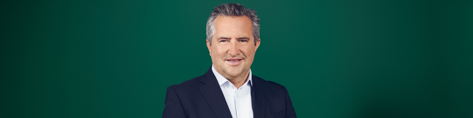 Rainer Schnabl, in dunklem Anzug und weißem Hemd, posiert vor einem einfarbig grünen Hintergrund. Mit einem leichten Lächeln blickt er in die Kamera und wirkt professionell und zugänglich. Das Bild ist klar und minimalistisch und konzentriert sich auf den Oberkörper und den Gesichtsausdruck des Porträtierten. Der schlichte Hintergrund unterstreicht den seriösen und formellen Charakter des Porträts.