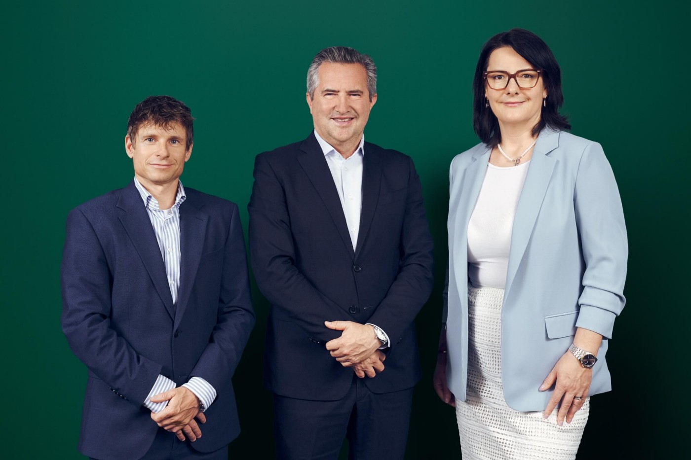 Gunter Deuber, Rainer Schnabl und Elitza Kavrakova stehen vor einem einfarbig grünen Hintergrund. Sie tragen formelle Geschäftskleidung, darunter Anzüge und ein Blazer mit Rock. Die zentrale Person steht mit verschränkten Händen da, während die anderen selbstbewusst zu ihren Seiten posieren. Das Bild vermittelt ein Gefühl von Unternehmenskultur, Führung und Teamarbeit.