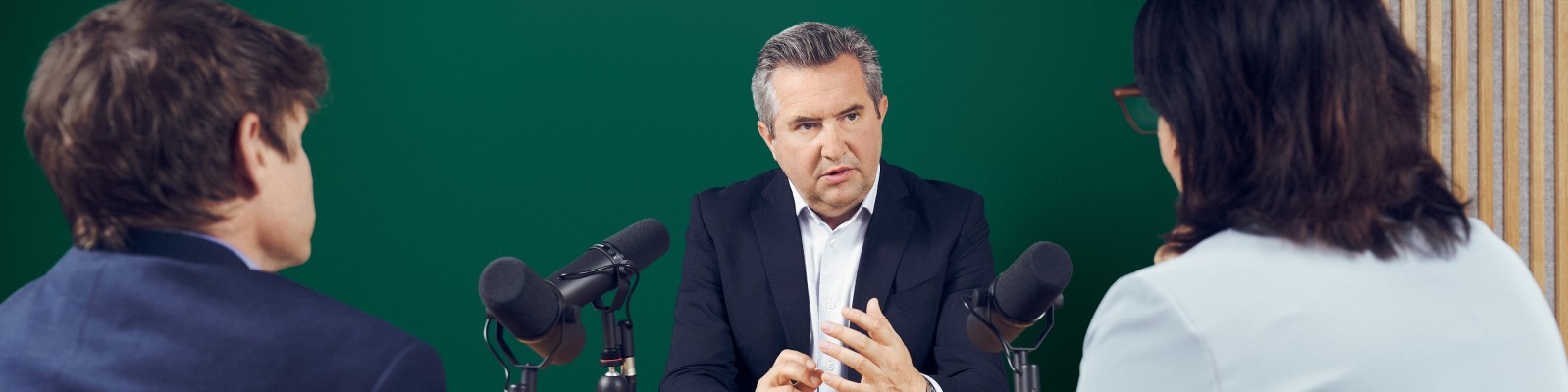 Rainer Schnabl sitzt im Anzug an einem Tisch und spricht während einer Interview- oder Podcast-Aufnahme in Mikrofone. Ihm gegenüber sitzen Elitza Kavrakova und Gunter Deuber in einem Studio mit grünem Hintergrund und Holzvertäfelung. Die Szene lässt auf ein professionelles Gespräch oder eine aufgezeichnete Geschäftsbesprechung schließen. Die klare und gleichmäßige Beleuchtung unterstreicht die formelle Atmosphäre.