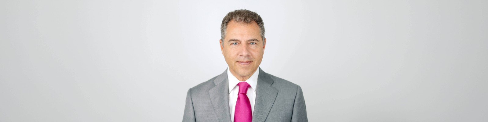 Axel Summer professionally dressed wearing a gray suit paired with a vibrant pink tie. The background is a plain, light-colored surface, emphasizing the formal attire. The image conveys a polished and business-oriented aesthetic.