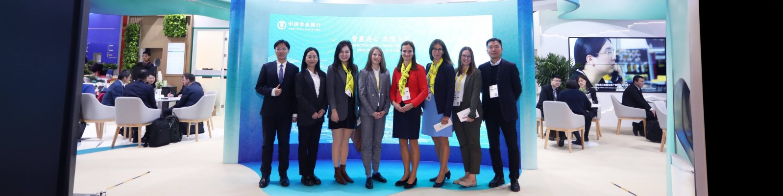 Eine Gruppe von neun Berufstätigen in Business-Kleidung, die zusammen vor einem blauen Display bei einer Konferenz oder Ausstellung stehen. Der Hintergrund zeigt eine moderne, gut beleuchtete Umgebung mit anderen Teilnehmern und Bildschirmen.