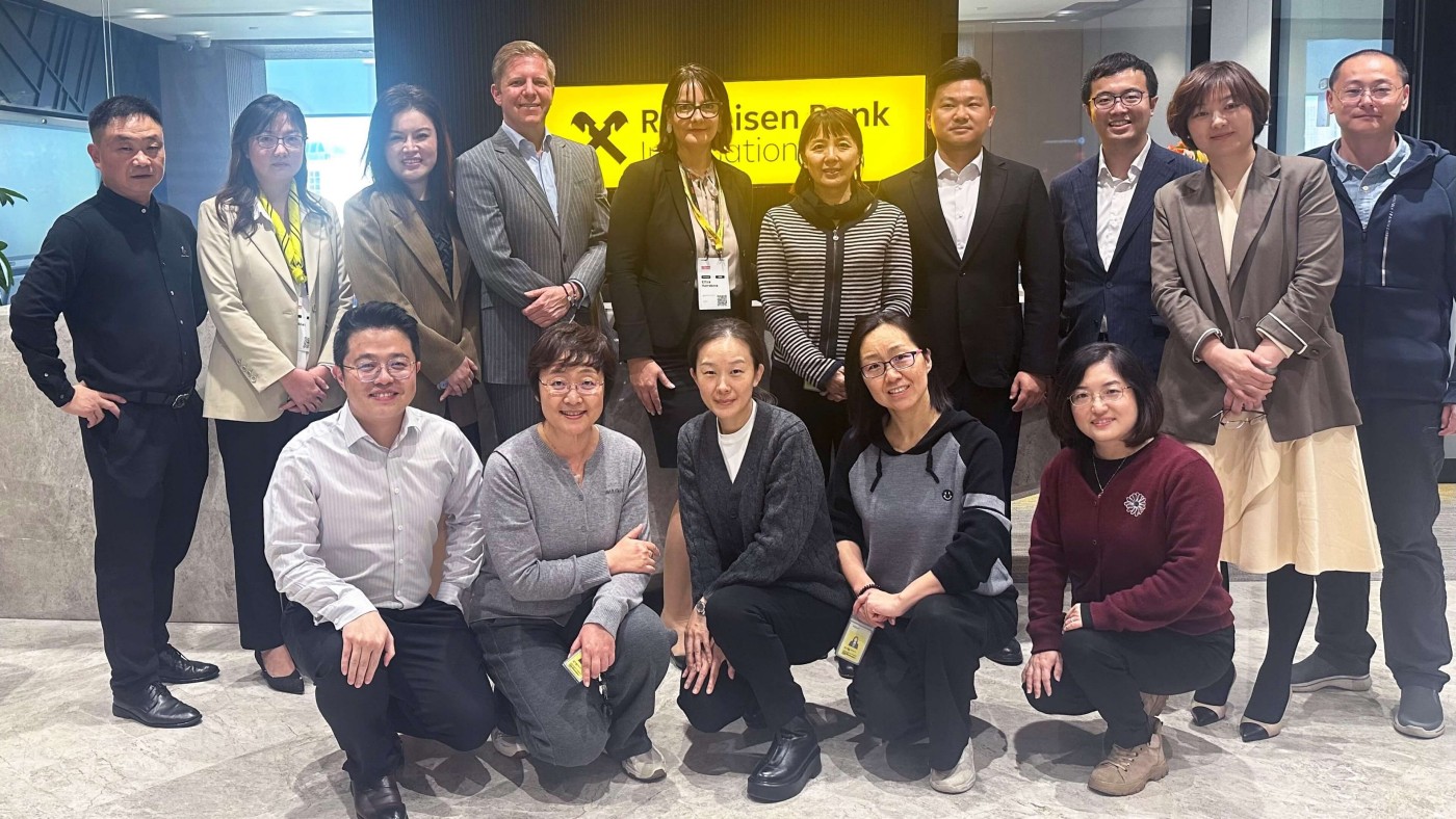 Eine Gruppe von Personen posiert gemeinsam für ein Teamfoto in einer Bürolobby mit einem gelben Raiffeisen Bank International Schild im Hintergrund.