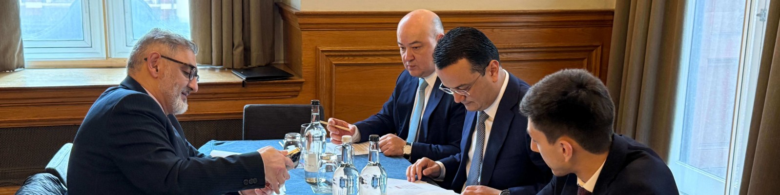 A group of professionals engaged in a business meeting around a table. Documents and water bottles are visible, indicating a formal discussion setting.